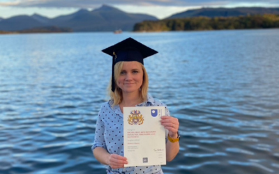 OU graduate Rebecca is standing, wearing a mortarboard and holding a degree certificate, with water and mountains in the background