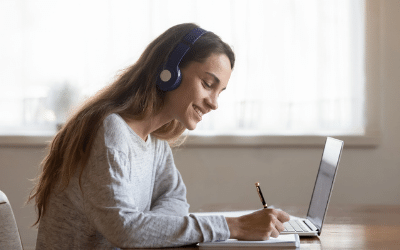 A smiling female is seated with a laptop, wearing headphones and writing in a notepad