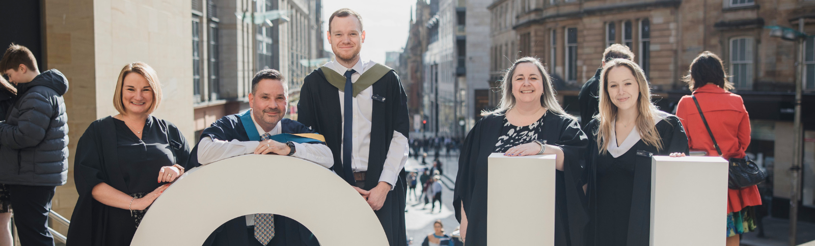 Photo of five people wearing graduation robes, standing in a line behind the giant white letters OU. 