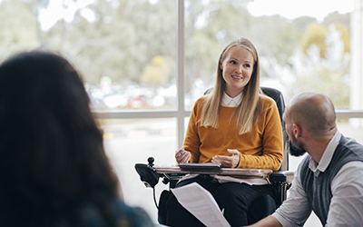 A woman sitting in a wheelchair, speaking to a man who is holding papers