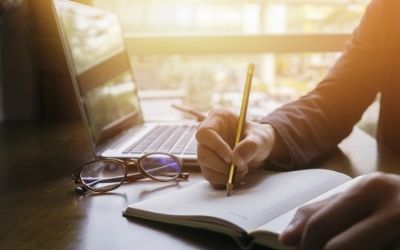 Image of a hand writing in a notebook with a pencil, beside an open laptop. 