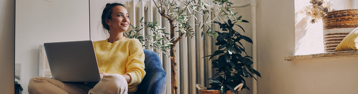 A woman sitting in armchair with her laptop, whilst smiling and looking out the window.