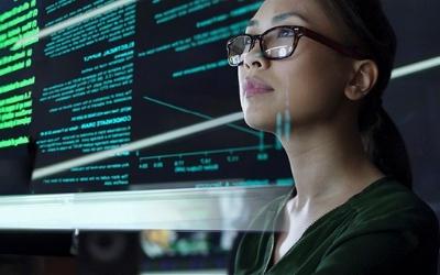 Photo of a woman looking at a computer screen, with computer screen text behind her too. 