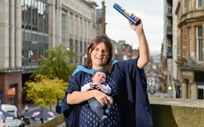 OU graduate Nicola Hehir wearing an OU graduation gown and holding her baby. 