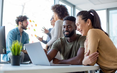 Two people seated and looking at a laptop screen, with three other people nearby who are in discussion. 