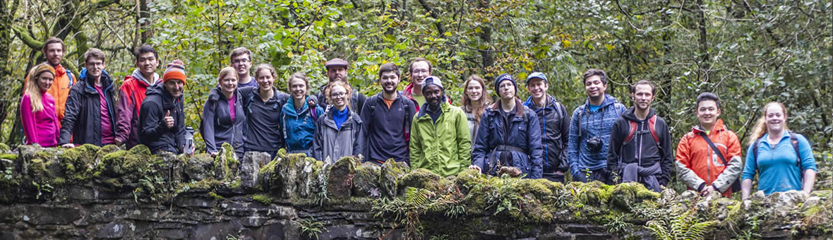 A large group in outdoor clothing stand for a photo on a bridge with trees in the background.