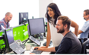 Two mature students looking at a computer screen. Two other students are in the background working on computers.