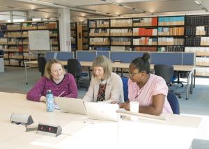 A group of colleagues are sitting together looking an open laptops. Behind them are rows of bookshelves.