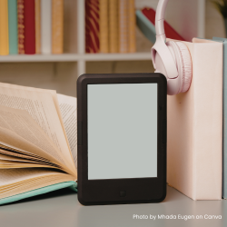 An open book, Kindle and headphones are placed together on a table, in front of a bookshelf. Photo credit: Photo by Mhada Eugen on Canva