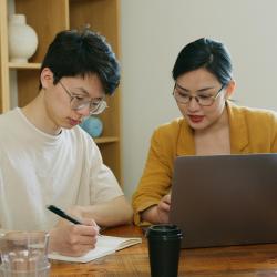 Two researchers sitting at a desk in conversation whilst typing on a laptop and writing in a notepad