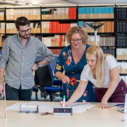 Three colleagues collaborate over documents on a large table in front of shelves of journals behind them.
