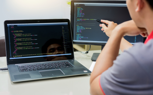 Person yn codio ar ddau liniadur